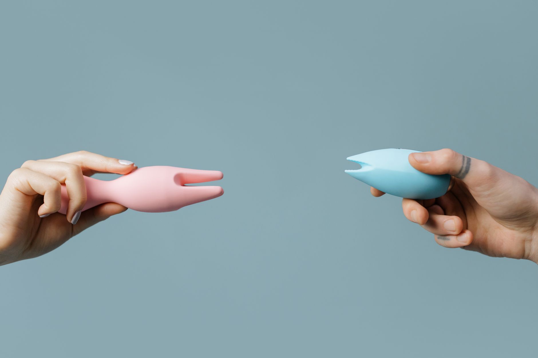 Two hands holding pink and blue silicone clitoral vibrators against a soft pastel background.