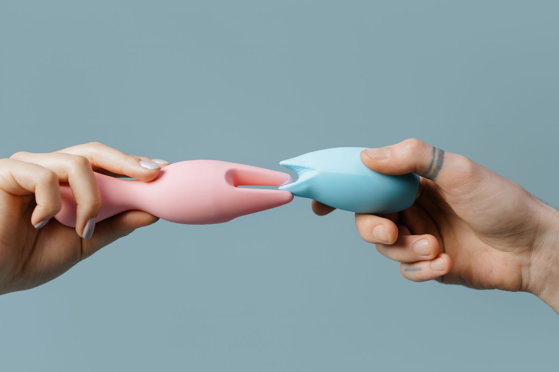Close-up of two hands holding pink and blue silicone clitoral vibrators against a soft pastel background